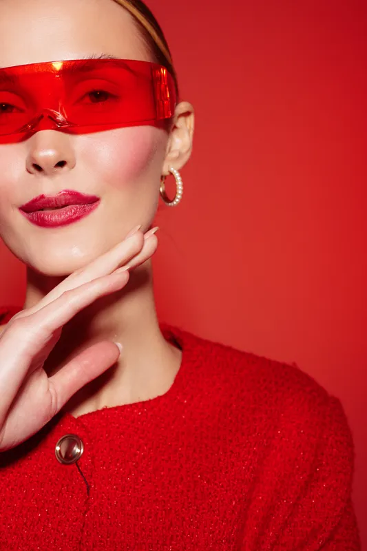 Close-up portrait of a stylish woman wearing red sunglasses and red outfit, posing with hand near face against a vibrant red background. Fashion and beauty concept.