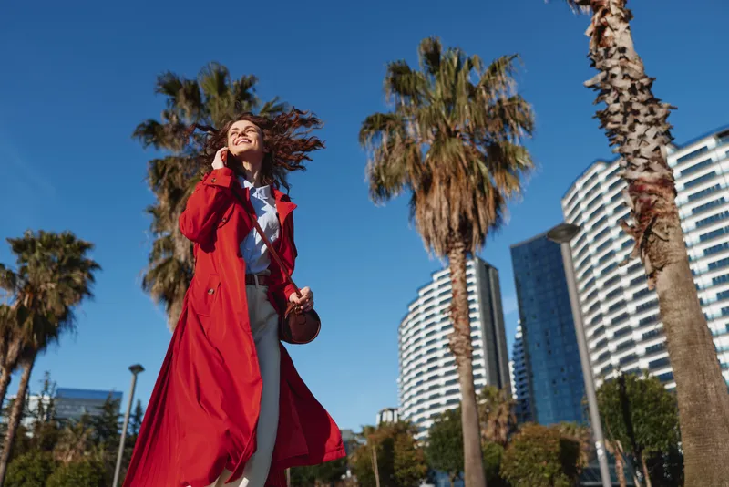 Vibrant woman in a red trench walks outdoors under palm trees, enjoying a sunny city moment with a confident stride and windswept hair, bright skies and modern towers framing her