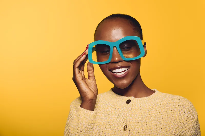 Smiling African woman with dark skin wearing large blue sunglasses and a beige knitted sweater on yellow background. Concept of fashion, summer style, and joyful mood.