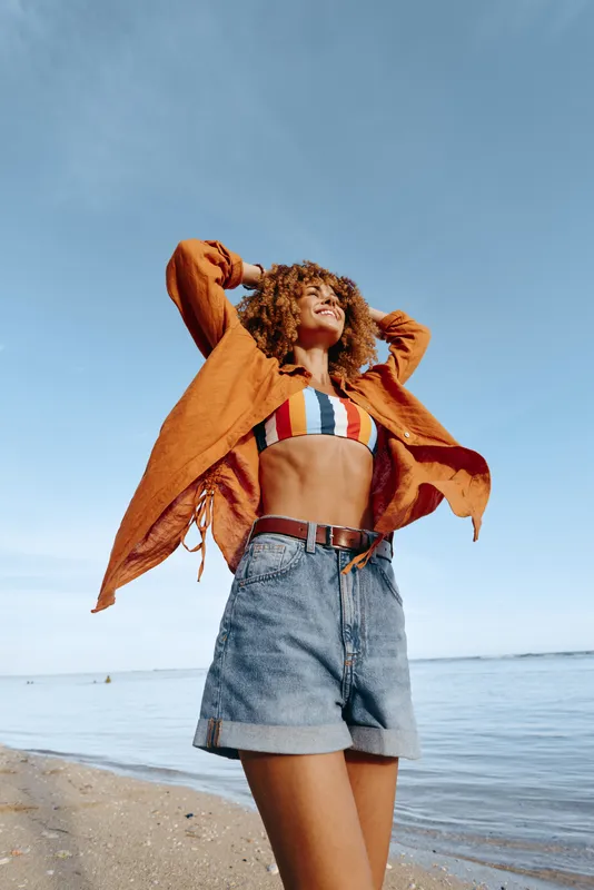 Young woman on a sunny beach wears a striped bikini top and denim shorts, lifting an orange jacket as the breeze blows, conveying carefree summer energy and confidence.