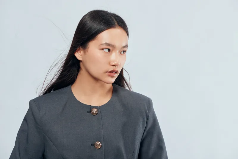 Young Asian woman with long dark hair wearing gray blazer looking aside on gray background. Studio portrait of businesswoman with natural makeup and serious expression.