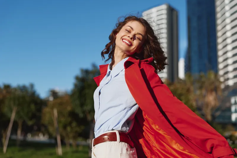 Young woman in a white shirt and red cape smiles as the cape billows in the wind against a sunlit city park backdrop with tall buildings nearby.