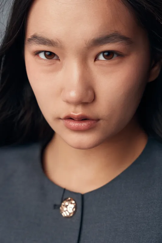 Young woman portrait with natural makeup and dark hair closeup. Asian female model with clear skin wearing gray top with decorative button in studio lighting.
