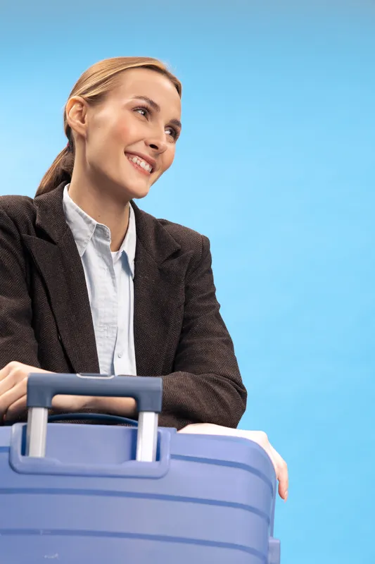 Smiling businesswoman in brown jacket with blue suitcase on blue background looks away happily. Concept of travel, business trip, vacation and positive lifestyle with luggage.