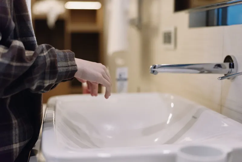 Person reaching toward a white bathroom sink with a chrome faucet, wearing a plaid sleeve, capturing a everyday moment of hygiene and hand washing in a modern restroom
