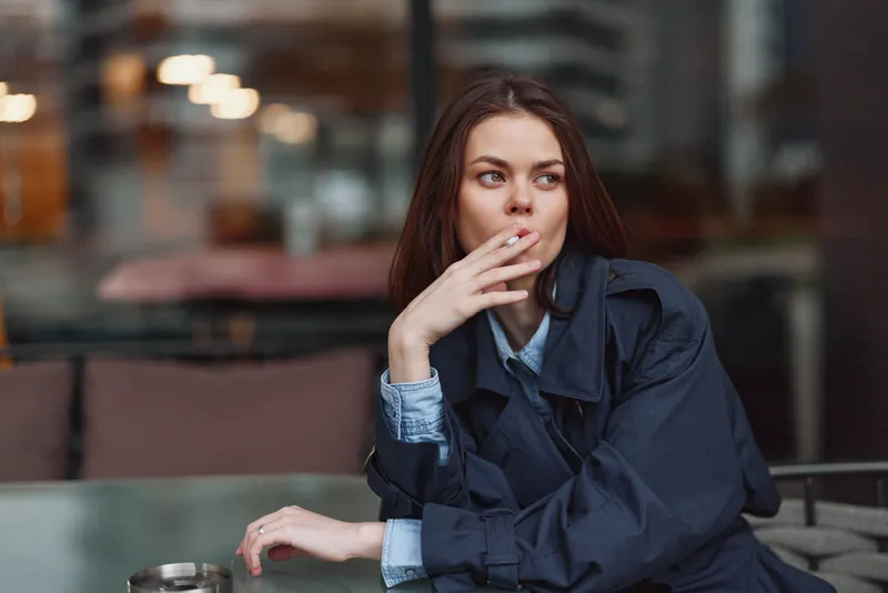 Young woman sitting outdoors in a dark coat, smoking cigarette, thoughtful expression, casual pose, blurred urban background, evening light