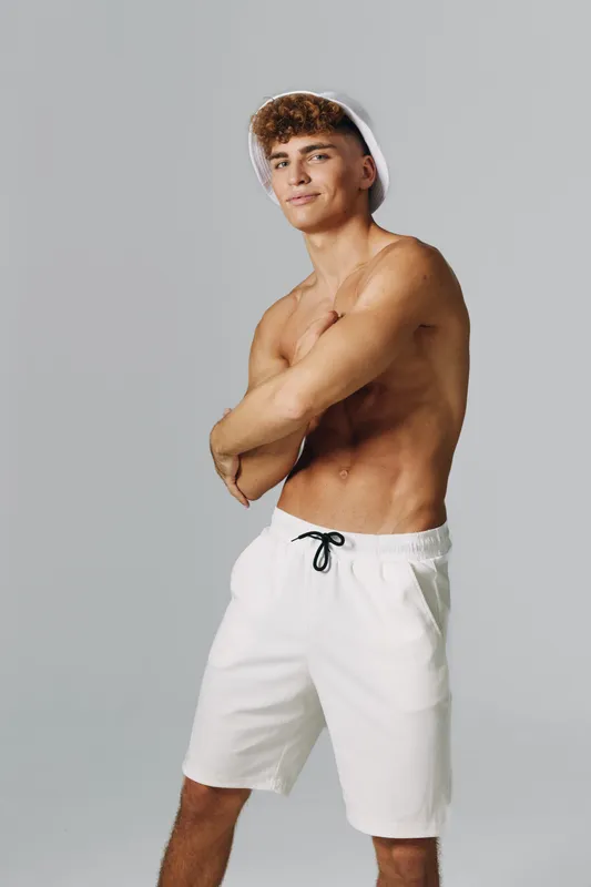 Young man standing shirtless with crossed arms wearing a white hat and white shorts on plain background. Confident model with athletic build posing for summer fashion concept.