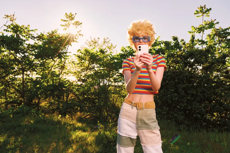 A retro outdoors portrait shows a fashionable model in striped shirt and shorts, taking a candid selfie in bright daylight, conveying nostalgic vintage vibes.