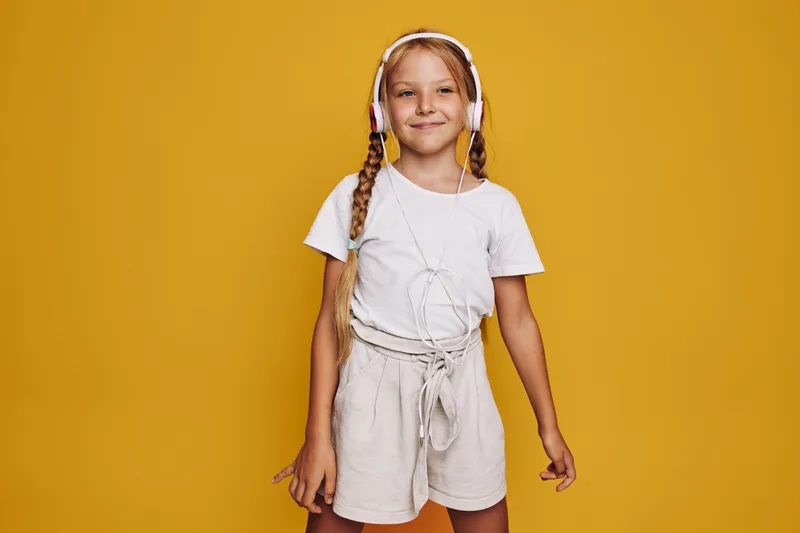 Young girl with braided hair wearing white headphones and casual outfit stands confidently against a yellow background. Concept for music, childhood, and happiness.