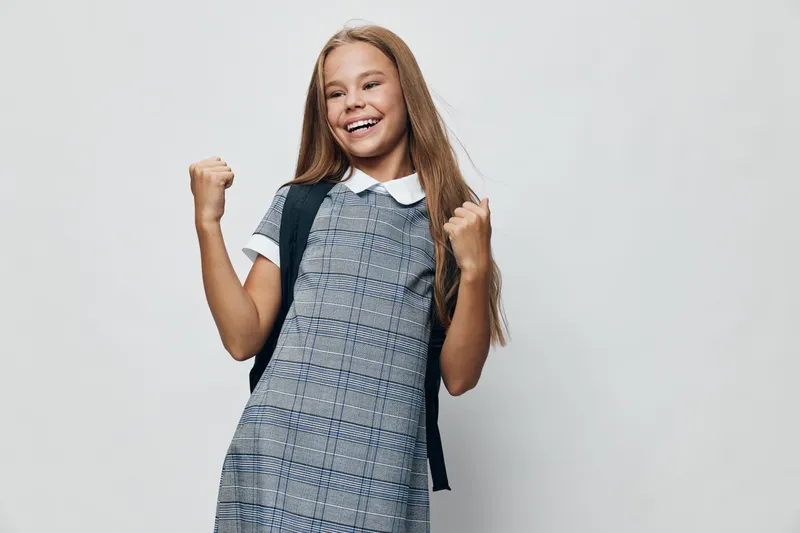 Girl in school dress with backpack smiling and clenching fists standing on white background for education, success and happiness in childhood and student life.