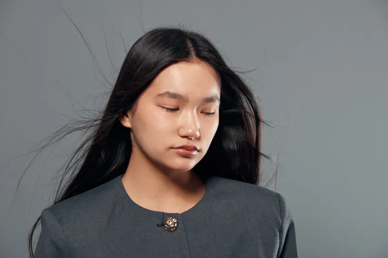 Young woman with long black hair wearing gray jacket with closed eyes in studio, portrait of calm female with natural makeup on neutral background, beauty and serenity concept.