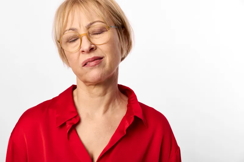 Senior blonde woman with glasses in red shirt portrait, eyes closed expression, calm peaceful mood, minimal white background, studio lighting, copy space.