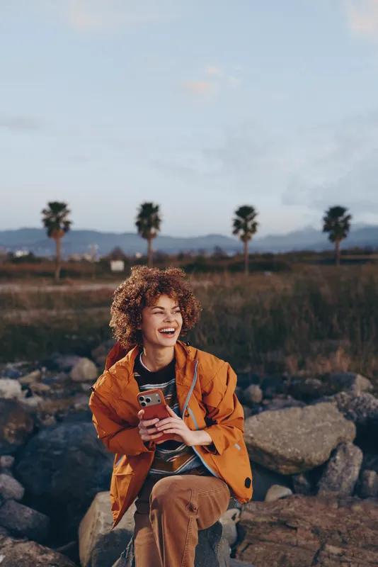 Woman lifestyle outdoors, orange jacket, smartphone in hands, smiling near rocks with palm trees and soft sunset sky, travel mood concept, casual lifestyle photo for modern communication