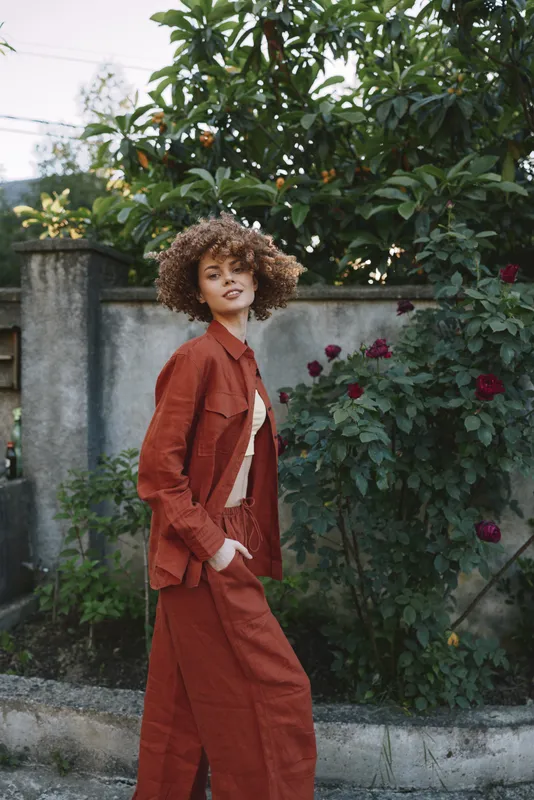 A woman in a rust suit stands outdoors, with lush greenery in the background, showcasing casual fashion and confident style as a lifestyle moment frozen in motion