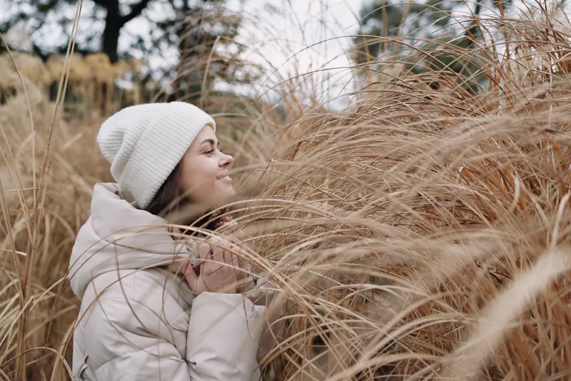 Woman in a winter coat and beanie enjoying tall grass in an open field, calm expression, soft light, outdoor nature portrait with chilly mood and seasonal scenery for lifestyle