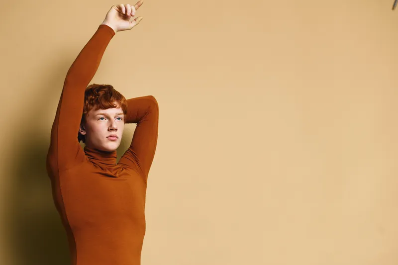 Young man with red hair wearing brown turtleneck sweater posing with hands behind head against beige background for fashion and casual portrait photoshoot.
