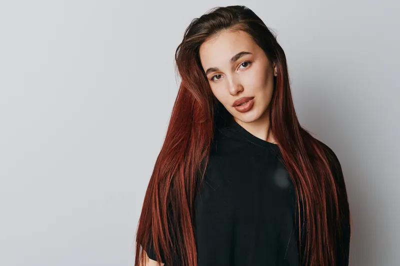 Young woman with long red hair and natural makeup posing in black shirt against plain background. Portrait with confident expression and casual style in studio lighting.