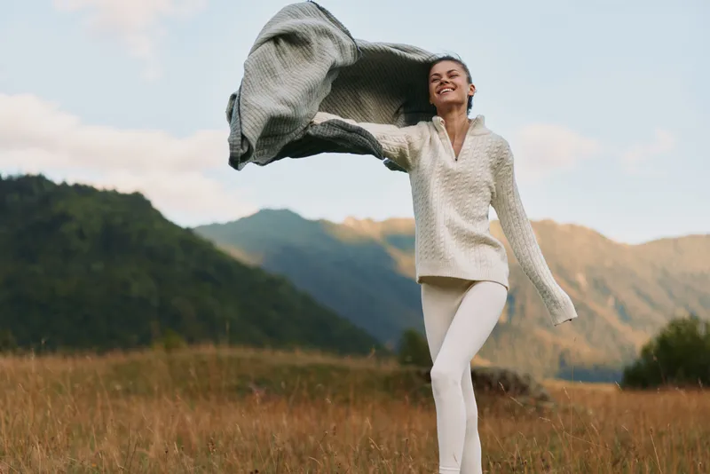 Young woman in white pants and knit sweater joyfully throws a gray scarf while standing in a grassy field with mountains in the background under a cloudy sky.