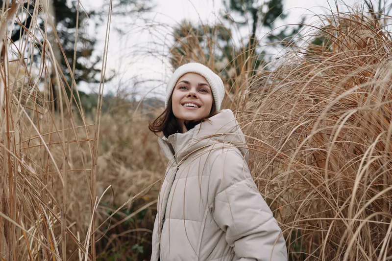 Woman in a light beige puffer coat outdoors amid tall grasses, smiling warmly as she enjoys a calm nature moment.