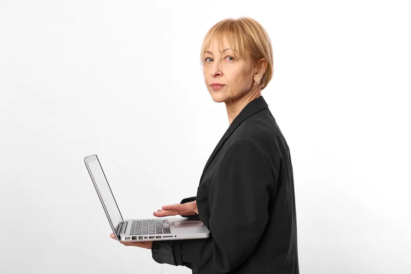 Older woman in business suit uses a laptop for portrait on a clean white background, calm focused expression. Professional communication and technology.