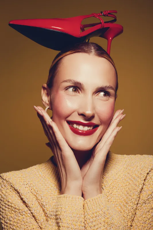 Smiling woman with red high heel shoe on head poses with hands near face. Studio portrait on brown background. Concept of fashion, creativity, and playful mood in beauty photography.