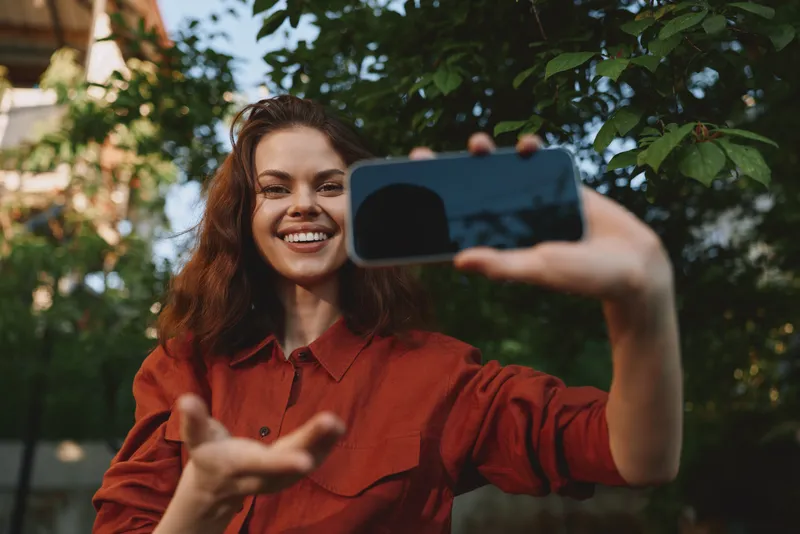 A smiling woman in a red shirt takes a selfie outdoors, capturing casual daily life and confidence. Bright natural light highlights candid lifestyle moments