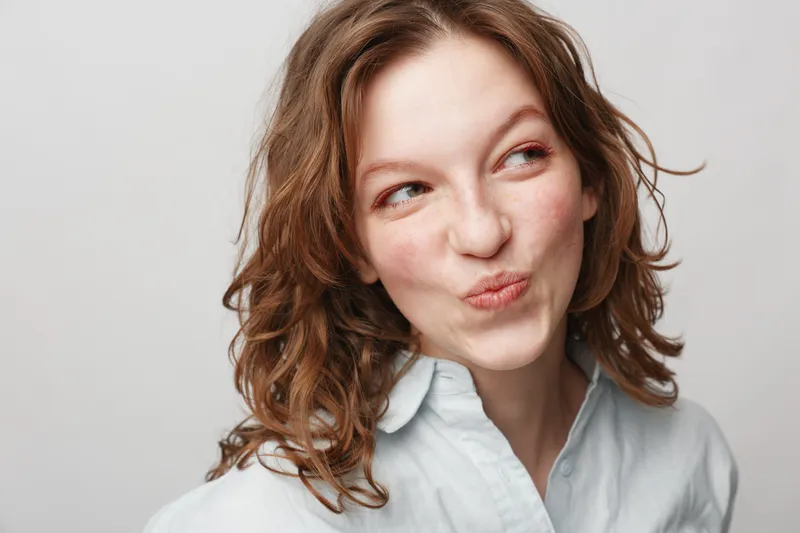 Young woman with curly hair wearing white shirt looks sideways with playful expression on light background. Concept of natural beauty, casual style, and candid emotions.