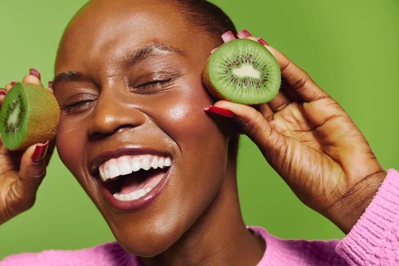Happy African woman with dark skin smiling with closed eyes holds two kiwi halves near face on green background. Concept of healthy lifestyle, fresh fruit and natural beauty.