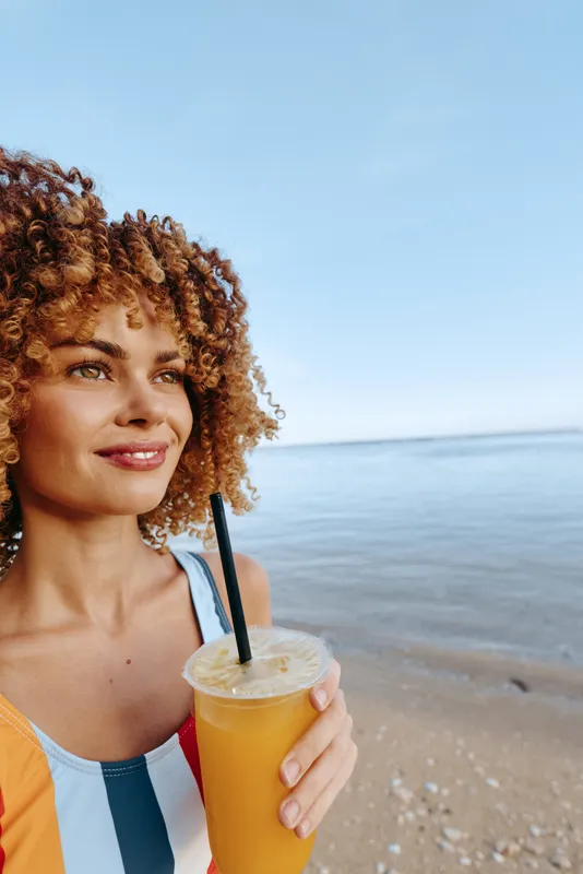 Woman with curly hair smiles on beach near ocean holding ice drink with straw, casual summer outfit, sunny daytime, sandy shore and calm horizon, close portrait outdoor