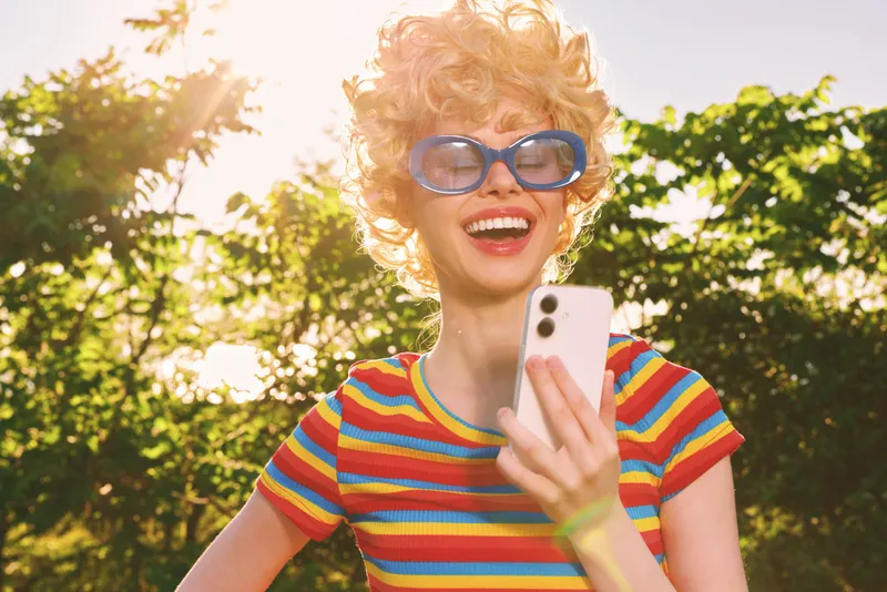Bright woman lifestyle shot with retro film color, outdoors in sun, wearing striped shirt and oversized sunglasses, smiling and using phone, playful candid moment.
