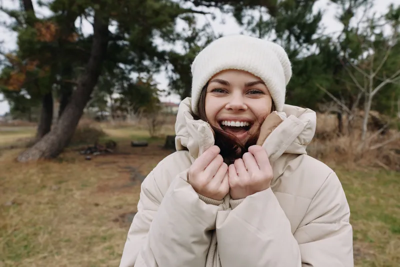 Woman outdoors wearing a warm winter coat and knit hat, smiling brightly in a park setting with trees and grass, casual cold weather moment.