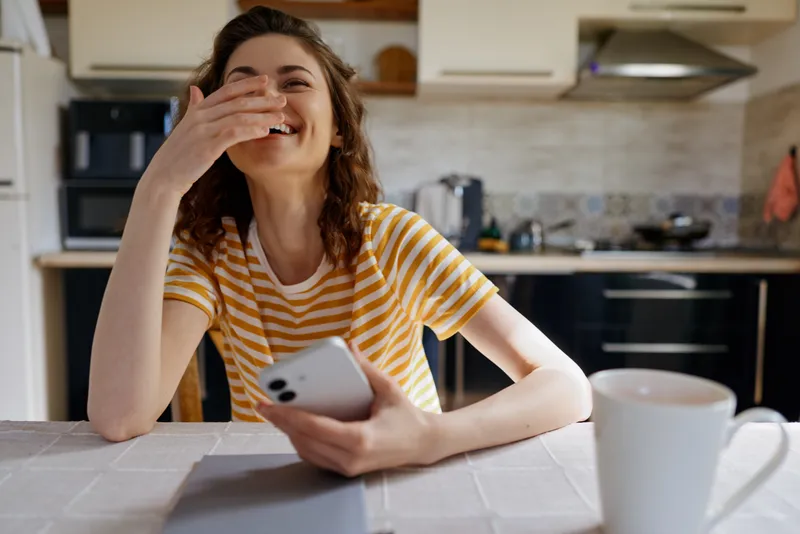 Woman in a yellow striped shirt sits at a kitchen table, laughing with her hand over her mouth, holding a smartphone, capturing a casual home lifestyle moment in a bright kitchen