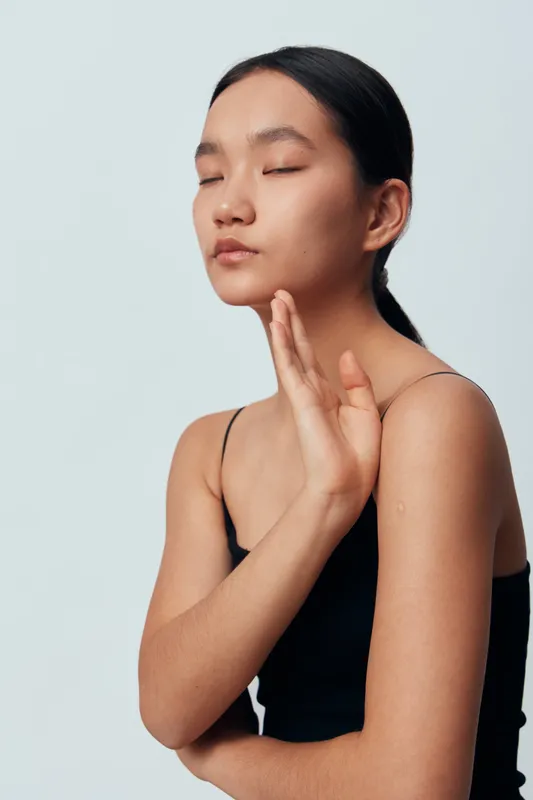 Woman with closed eyes and calm face in black sleeveless top posing with raised hand on light background. Beauty, serenity, relaxation, skin care, mindfulness and meditation concept portrait.