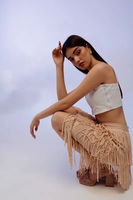 Young woman in fashion outfit poses against a plain background. She wears a white crop top and fringed beige skirt. Concept of beauty, style, and modern fashion.