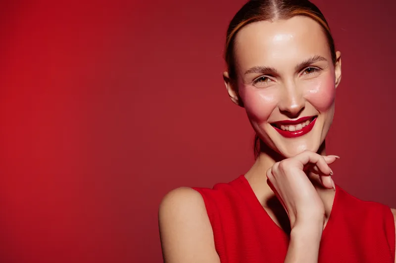 Smiling woman with red lipstick and blush on cheeks poses against red background wearing sleeveless red top. Concept of beauty, makeup, fashion, and confident expression.