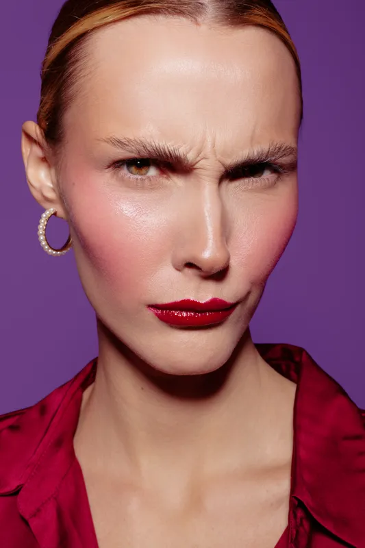 Close-up of a woman with furrowed brows and red lipstick wearing hoop earrings and a red blouse. Purple background highlights her intense facial expression and makeup details.
