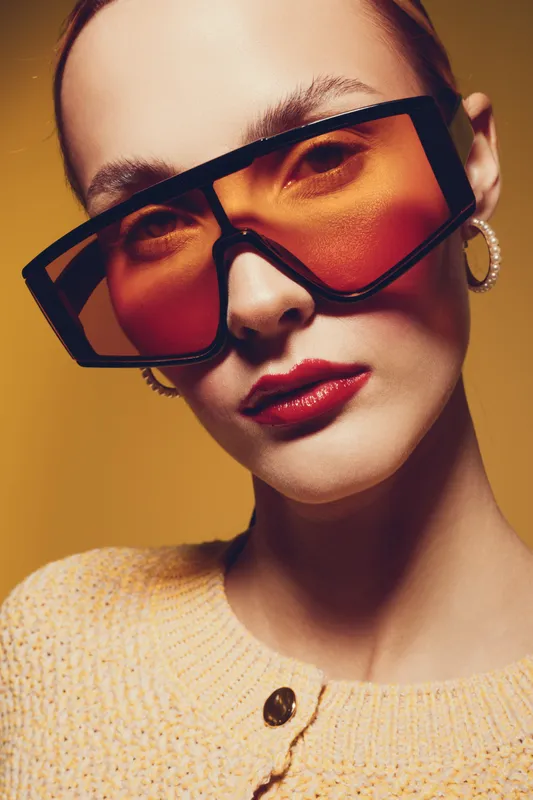 Close-up portrait of a fashionable woman wearing oversized orange sunglasses and hoop earrings. She has red lipstick and a beige knitted sweater on a warm yellow background. Concept of style and