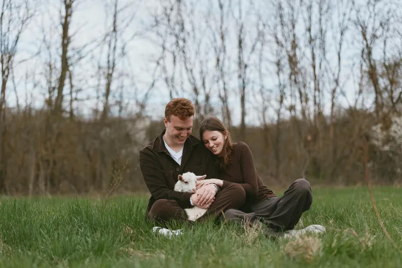 A couple with a goat kid relaxes outdoors in a grassy field, sharing a gentle, candid moment. This lifestyle scene highlights companionship, tenderness, and the simple joys of rural life.