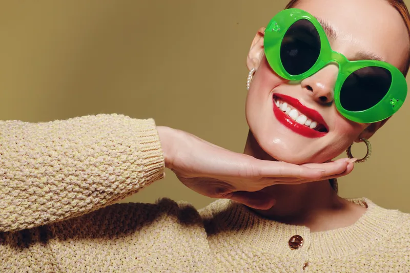 Smiling woman with bright red lipstick wears large green sunglasses and beige knitted sweater. She poses with hand under chin against plain background. Concept of fashion, style, and confidence.