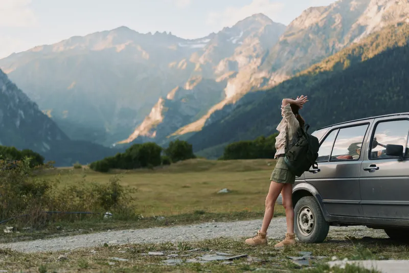 Young woman in casual clothes stretching beside a car on a mountain road. Scenic landscape with green fields and rocky peaks in the background. Concept of lifestyle and travel.
