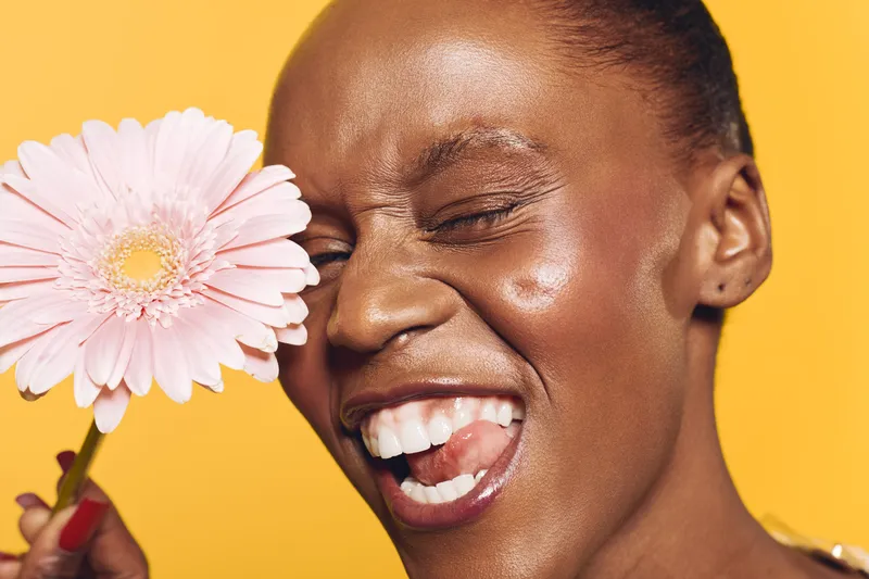 Happy smiling African woman with dark skin holding pink flower near face on yellow background. Close-up portrait with natural makeup, skincare concept, beauty and positive emotion.