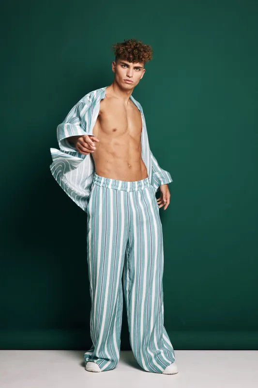 Young man with curly hair wearing striped pajama pants and open shirt posing against green background. Fashion model with toned body and casual sleepwear style in studio.
