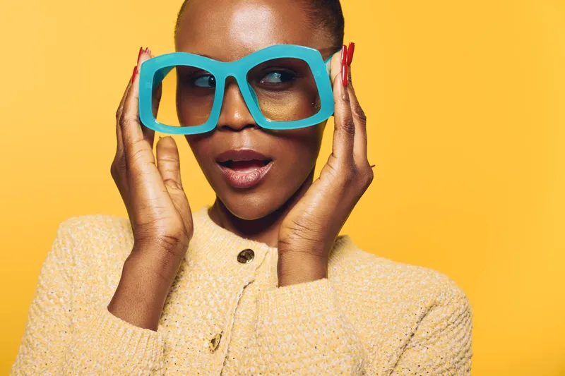Close-up of a fashionable black woman with dark skin wearing oversized blue glasses and a beige sweater. She poses against a bright yellow background, concept of style and confidence.