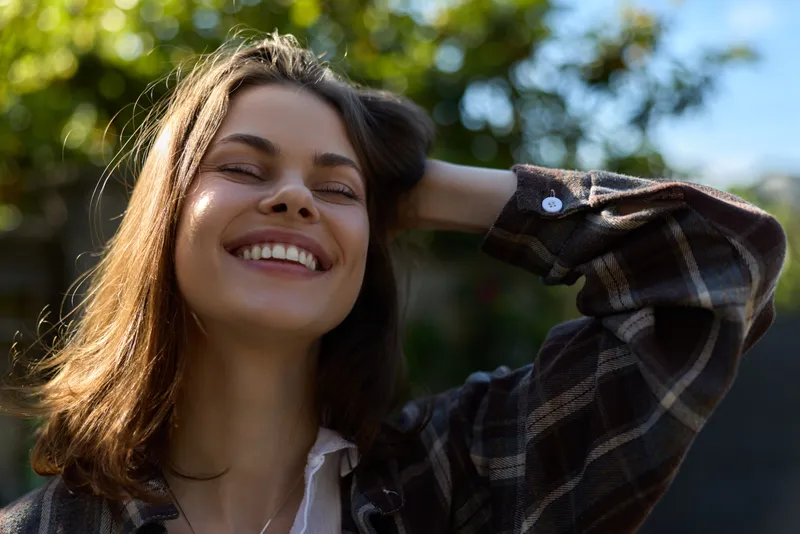 A smiling woman in casual wear enjoys a sunny outdoor moment, with one hand behind her head and eyes closed. Bright sunlight and leafy background convey carefree, everyday happiness