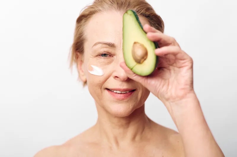 Smiling mature woman with natural skin holds half avocado near face while applying white cream on cheek. Concept for natural skincare, healthy aging, beauty routine, and organic wellness.