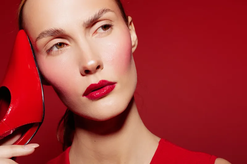Close-up of a woman with red lipstick holding a red high heel shoe near her face on a red background. Concept of fashion, beauty, and style with vibrant colors.