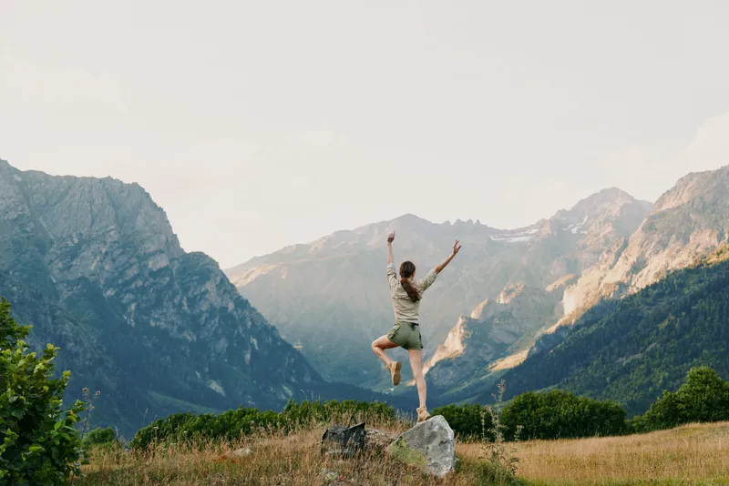 Young woman outdoors jumping on rock with arms raised, mountain landscape background, sunny day, casual outfit, active lifestyle, natural environment