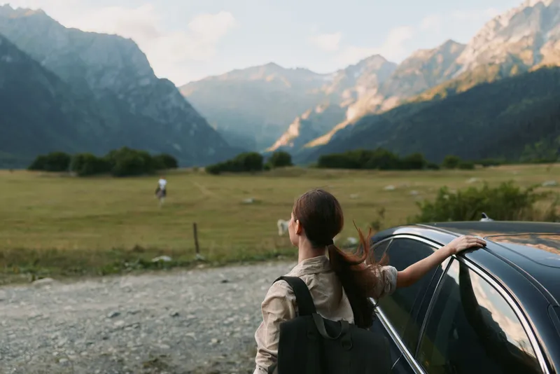 Young woman with long brown hair stands next to a black car looking at mountains in a wide open field during daytime.