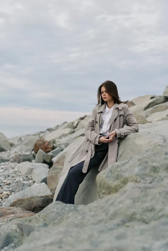 Young woman in trench coat sits on rocky shore under cloudy sky, looking down pensively. Casual autumn fashion concept with natural landscape and moody atmosphere by the sea.
