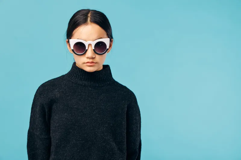 Young woman wearing white sunglasses and black turtleneck sweater standing against blue background, serious expression and dark hair tied back for casual fashion style.